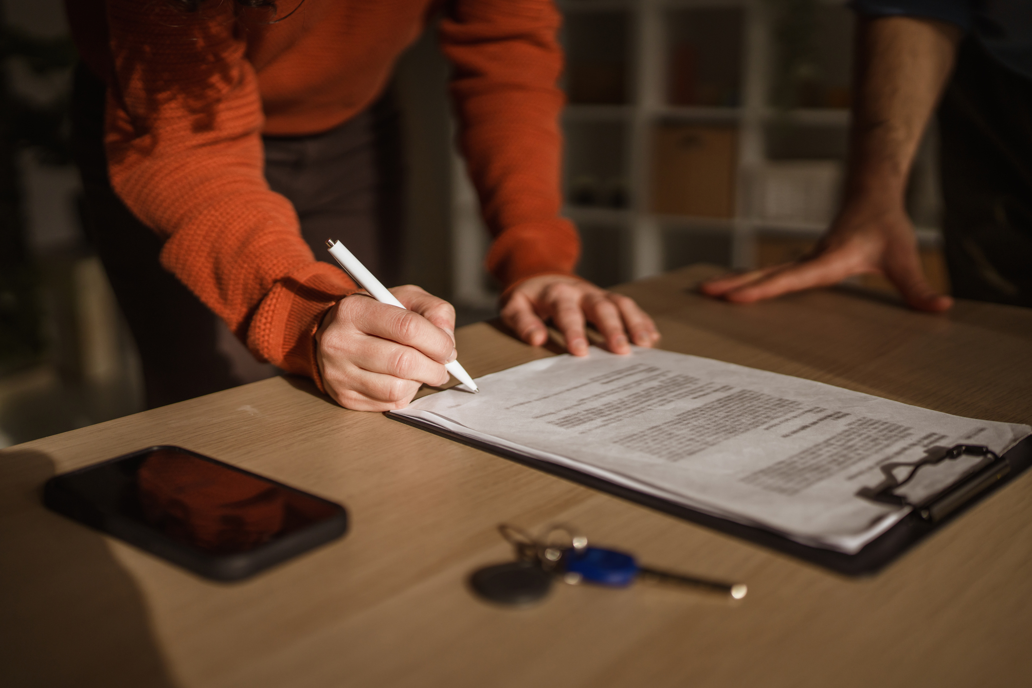 Woman's hand writing signature on a formal document during a property transaction, with car keys and a smart phone on wooden desk, symbolizing new ownership