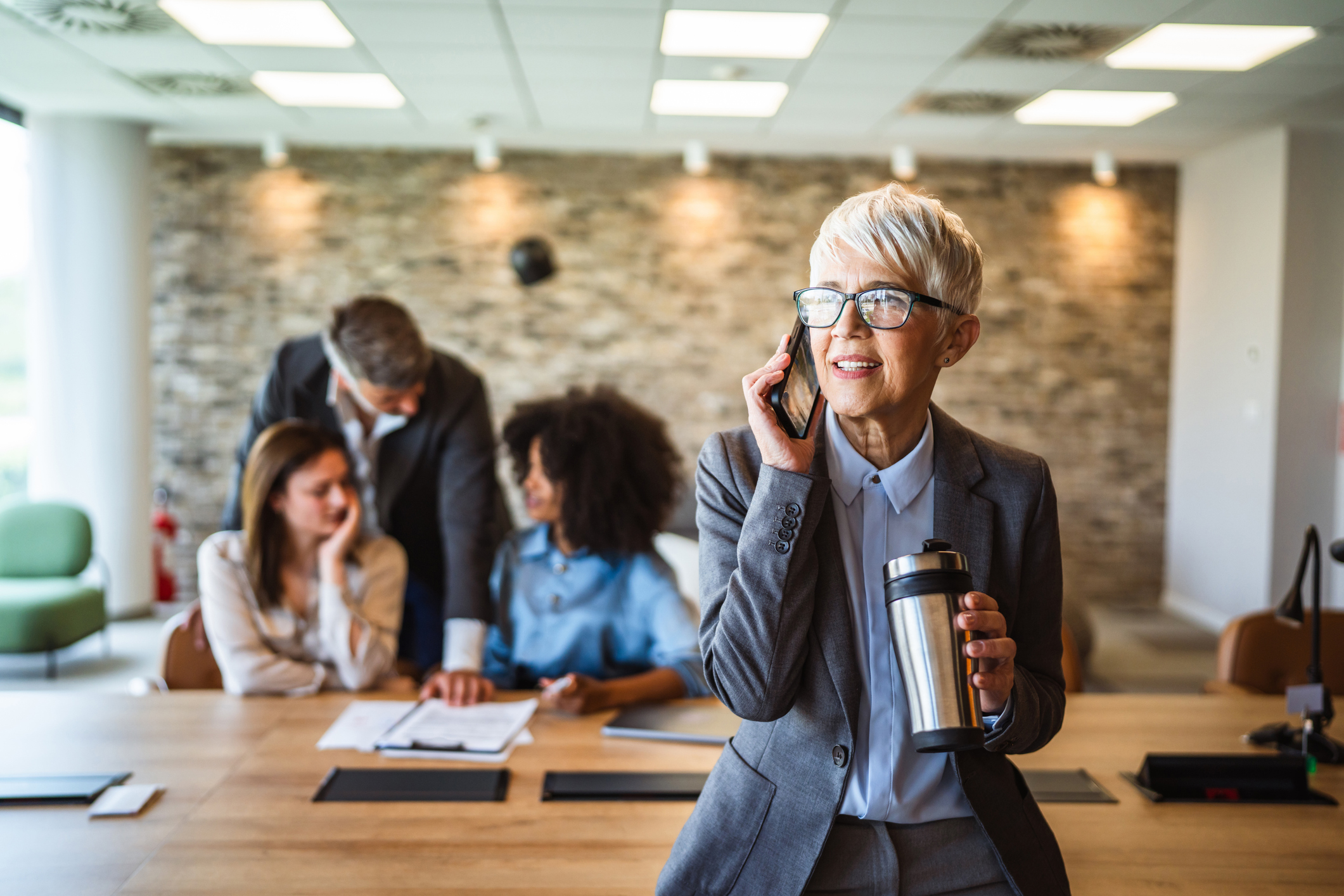 aged businesswoman talk on mobile phone and drink coffee at workplace
