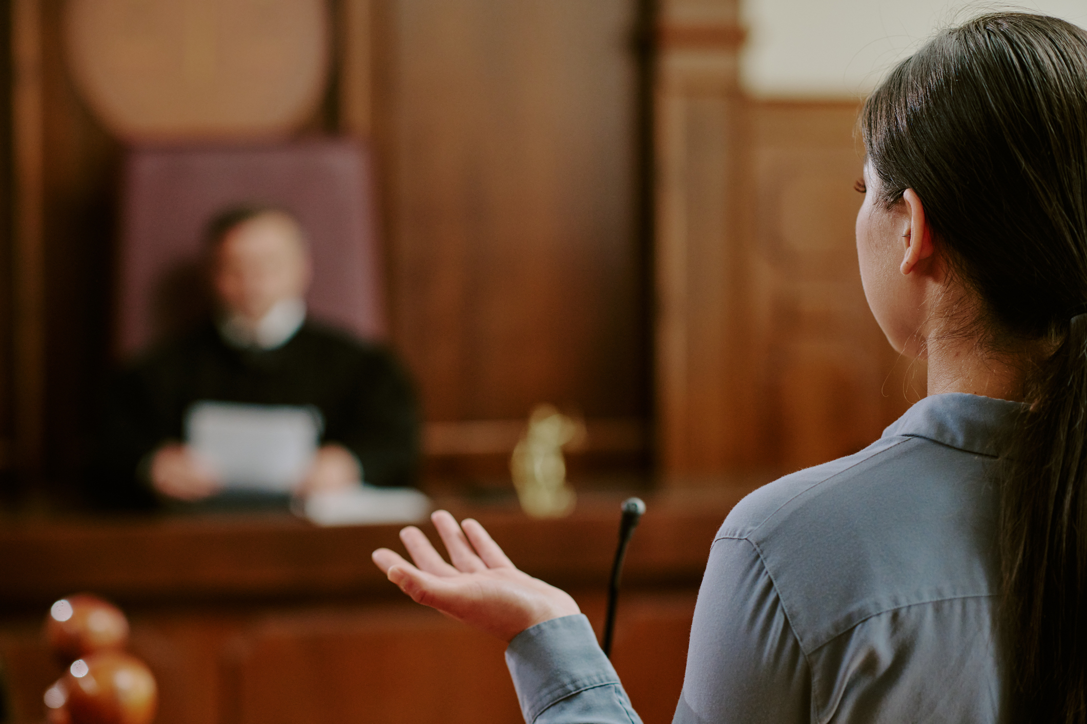 Young adult Asian woman testifying in courtroom, standing in front of judge, gesturing with hand while speaking, legal proceeding taking place in judicial setting